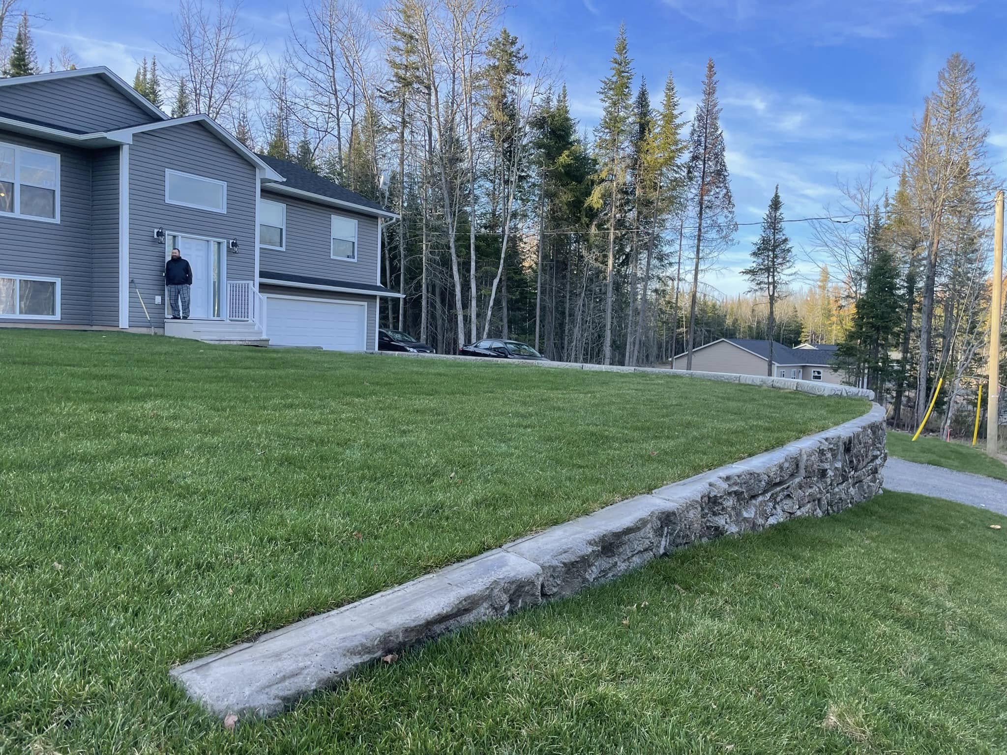 Two-story gray house with a curved stone retaining wall and a well-manicured lawn. A driveway and trees in the background.