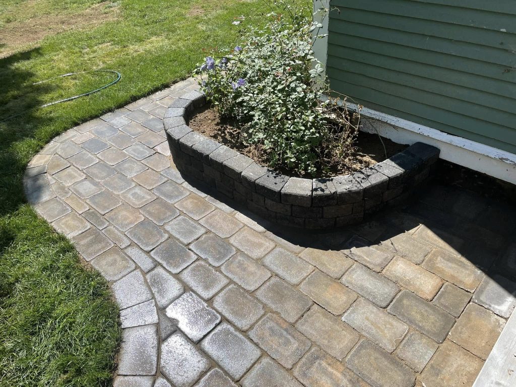 This image shows a small, curved brick pathway leading up to a house with light green siding. Adjacent to the path is a raised flower bed made of dark bricks, containing some plants and flowers.