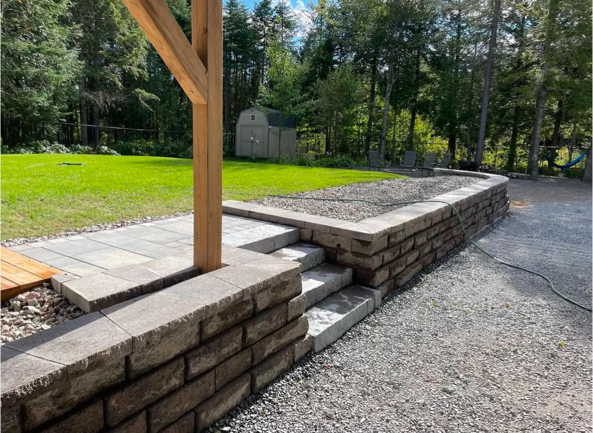 The image shows a partially completed outdoor patio or walkway project. The area features a raised brick retaining wall supporting a section of the paved or tiled surface.