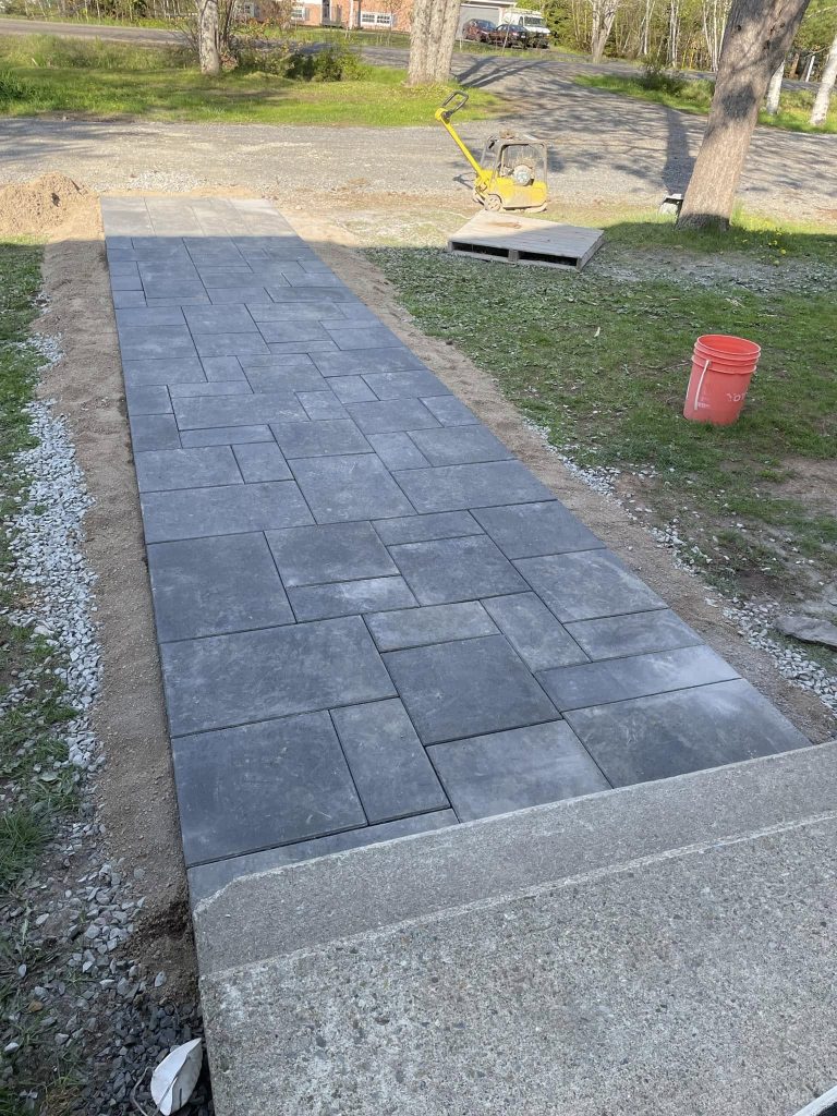 Long rectangular paved walkway/driveway with grey interlocking concrete pavers, flanked by dirt and gravel, with a red bucket and construction equipment in the background.