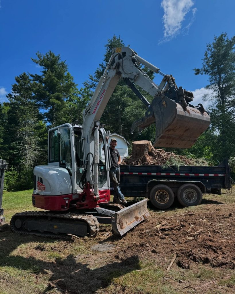 Mini excavator loading a pile of dirt/roots into a dump trailer, with a person standing on the trailer bed.
