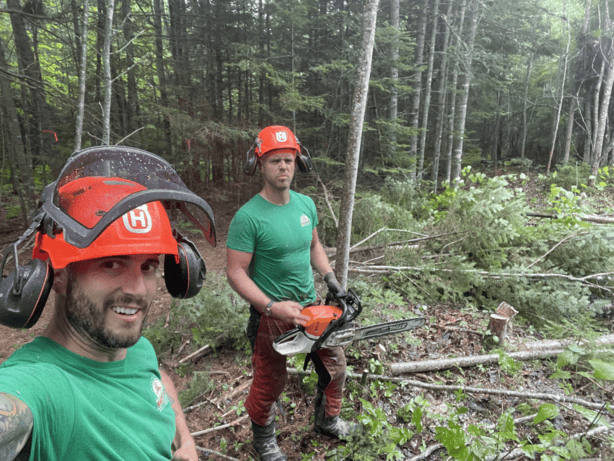 Two workers in green shirts and orange helmets in a forested area, one taking a selfie and the other holding a chainsaw near fallen branches and logs.